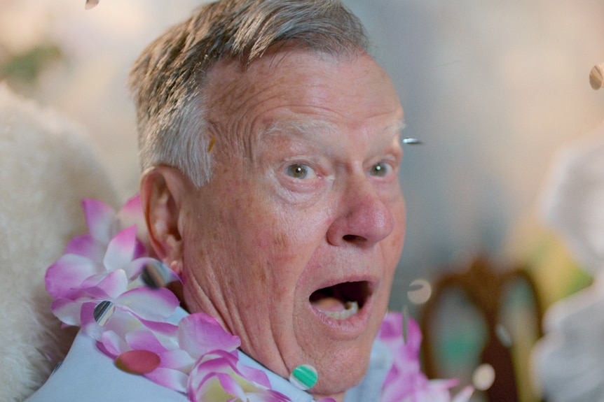 An older man sits with shocked and excited expression, greying hair wears pink and white Hawaiian flower lei around neck.