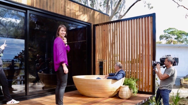 attractive woman stands on deck while tv host sits in wooden outdoor bath and they are filmed