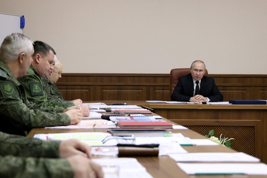 Vladimir Putin sits at the head of a table with people in defence uniforms sitting on the side. 
