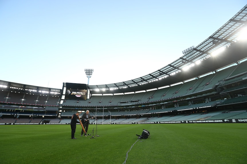 A man with a guitar and an Indigenous man with a didgeridoo play in an empty AFL ground, with a big screen in the background.