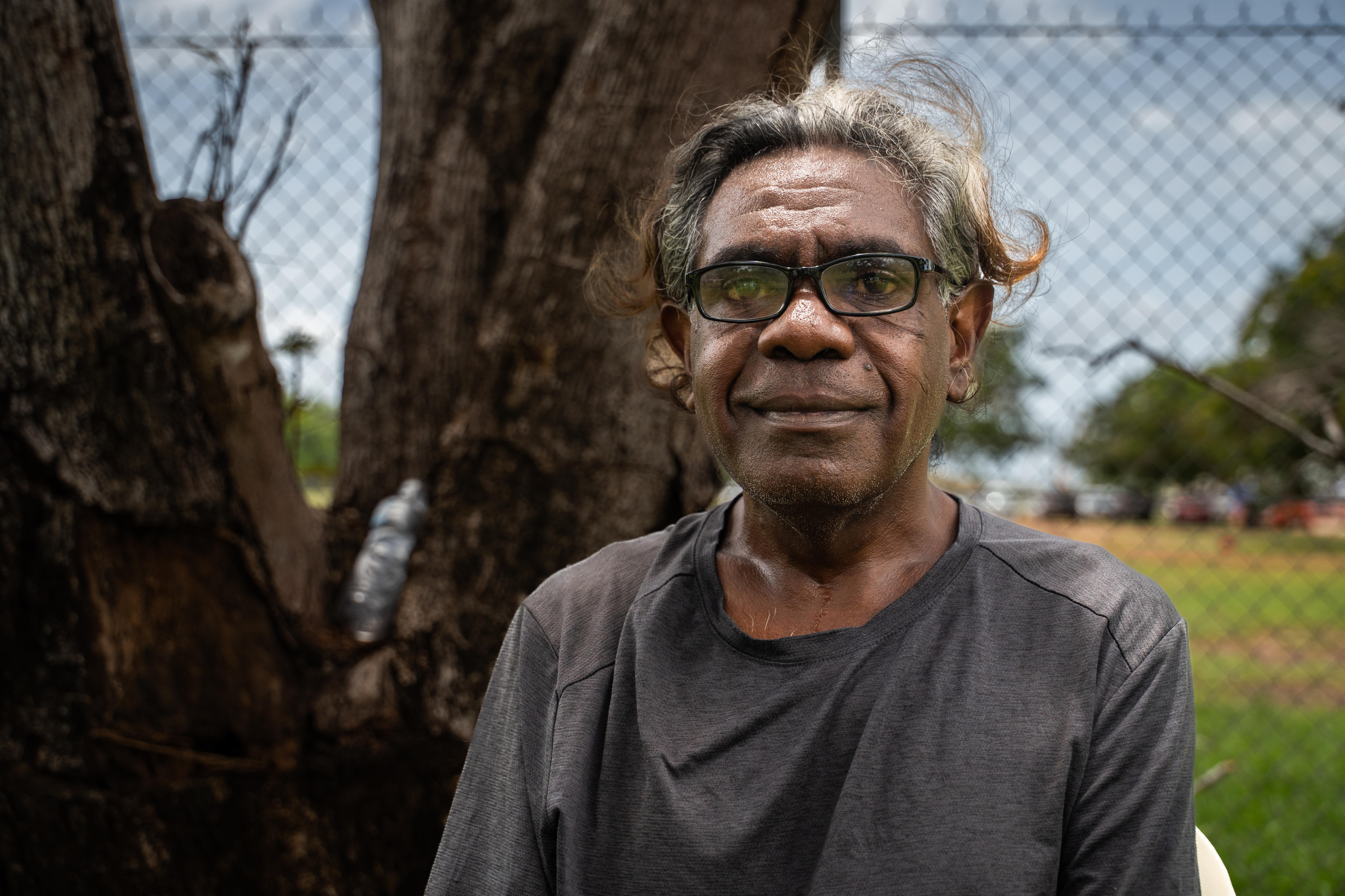 Un hombre de pelo corto y gafas oscuras sonríe levemente a la cámara. Detrás de él hay una alambrada.