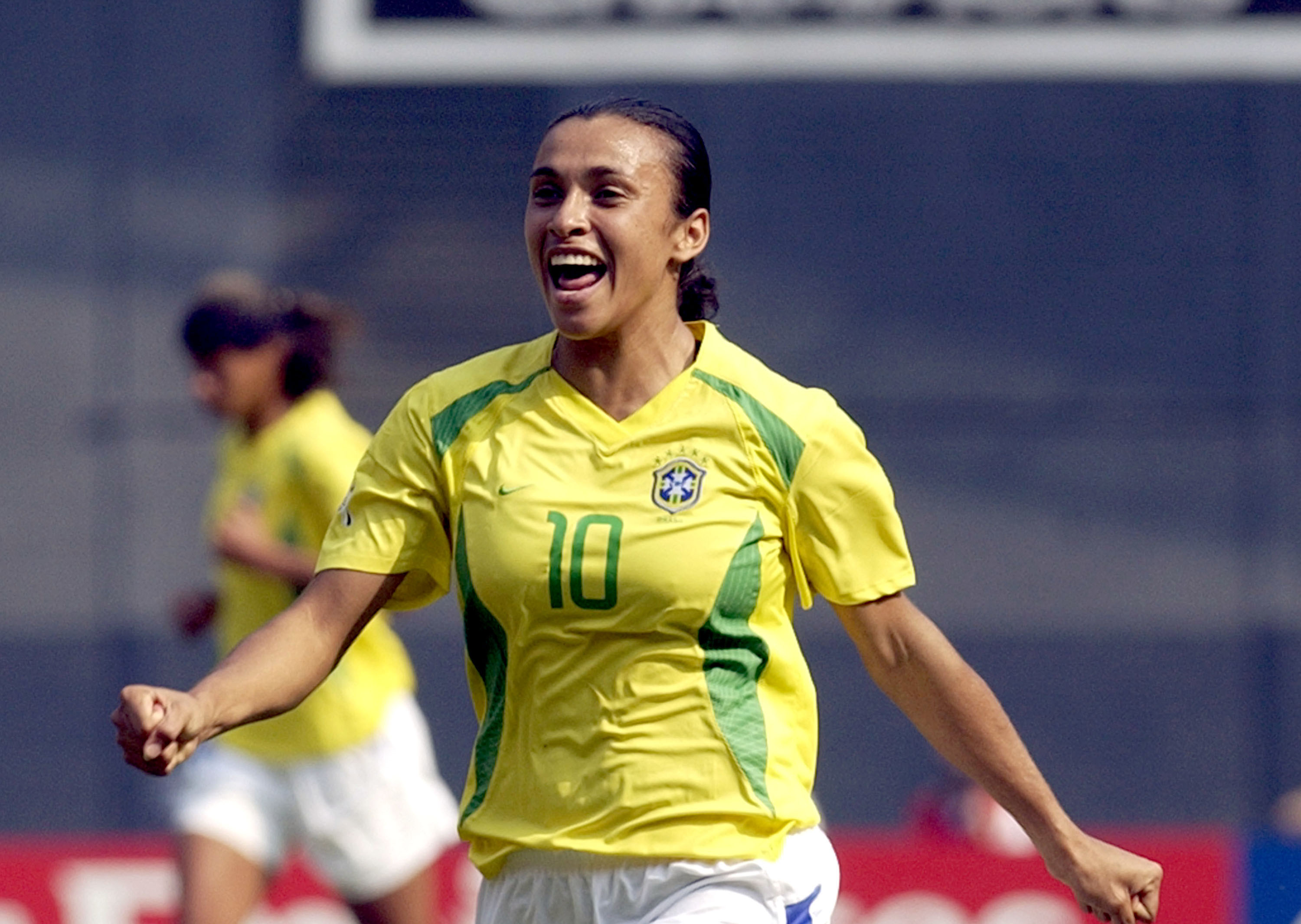 A young woman grins while running in a yellow and green soccer jersey.