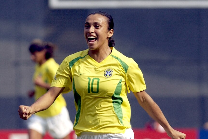 A young woman is smiling while running in a yellow and green soccer jersey after a goal is scored.