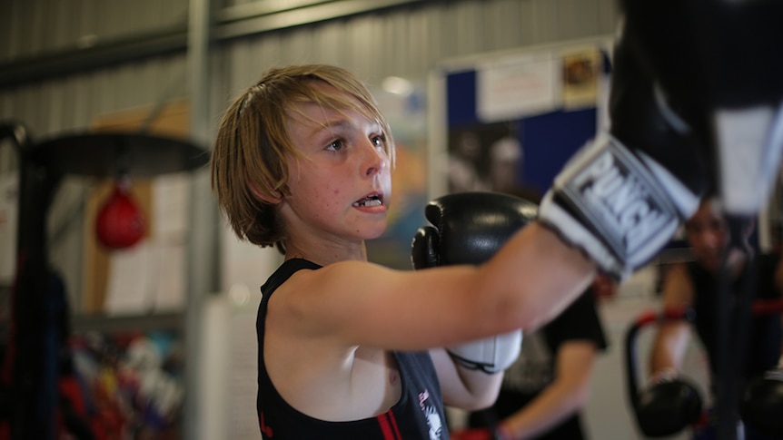 A young blonde boxer wears gloves as he hits the bags in a boxing gym.
