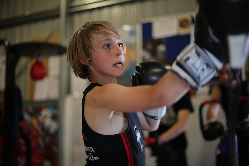 A young blonde boxer wears gloves as he hits the bags in a boxing gym.