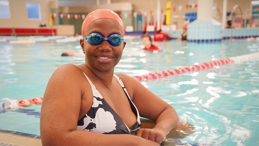 A woman wearing googles and a swimming cap sits in an indoor pool