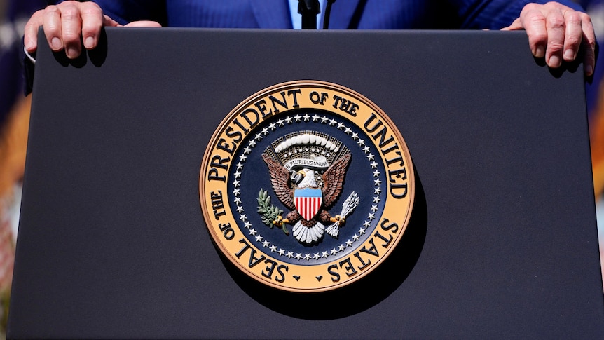 Two hands grip a lectern with a seal on the front saying SEAL OF THE PRESIDENT OF THE UNITED STATES