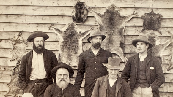 Bushmen and their dogs seated in front of a wall of animal skins (including koala pelts), circa 1870-1900.