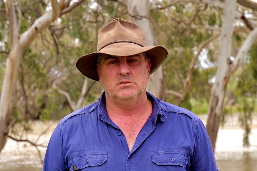 A man wearing a hat, blue shirt standing in front of the Darling River in Menindee
