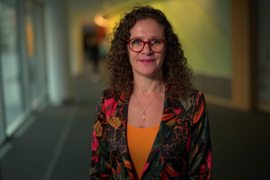 Sophie in 't Veld stands in a corridor in low-light, wearing red-rimmed glasses, with a small smile, looking into the camera