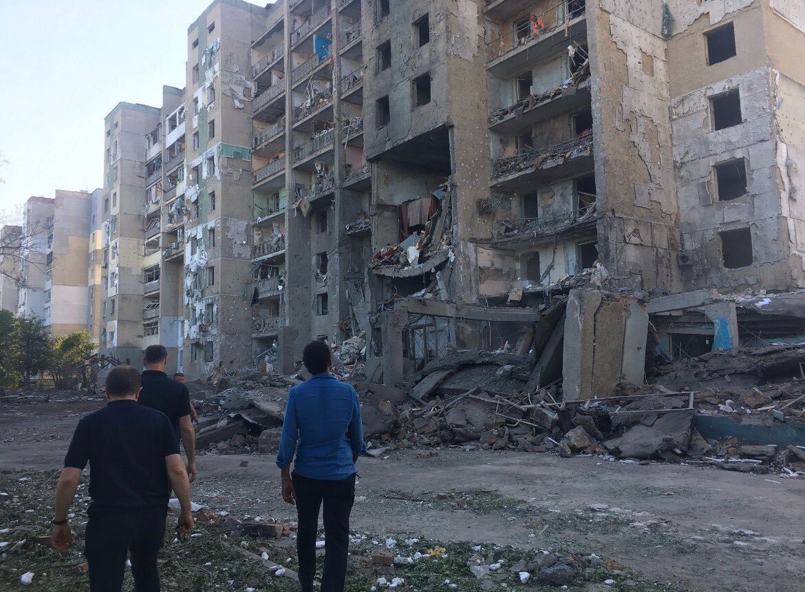 People standig in dirt look up towards a damaged building. 