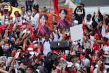 An animated crowd of protesters, many wearing red and white, hold posters, pictures and a speaker up as they demonstrate.