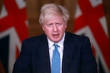 UK Prime Minister Boris Johnson furrows his brow. He wears a navy suit with blue tie and stands in front of two Union Jack flags
