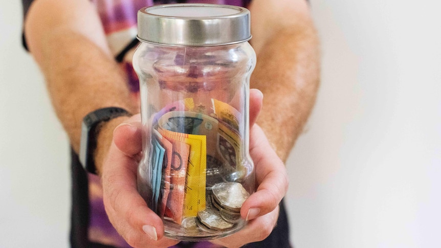 A male figure in a t-shirt holds out a clear glass jar half filled with Australian bank notes and coins.