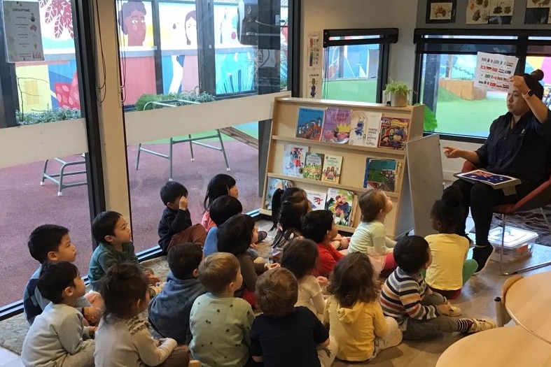 Dans une salle de classe colorée, de jeunes enfants sont assis par terre et fixent un enseignant, assis, en train de leur lire un livre.