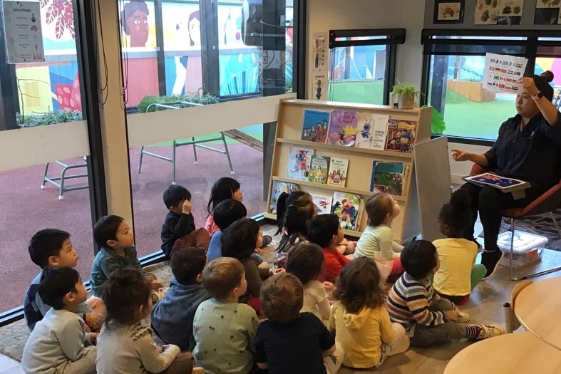 In a colourful classroom, young children sit on the floor staring up at a teacher, seated, reading a book to them.