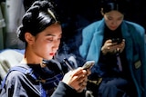 A Chinese woman with her hair braided looking at her phone while holding a vape 