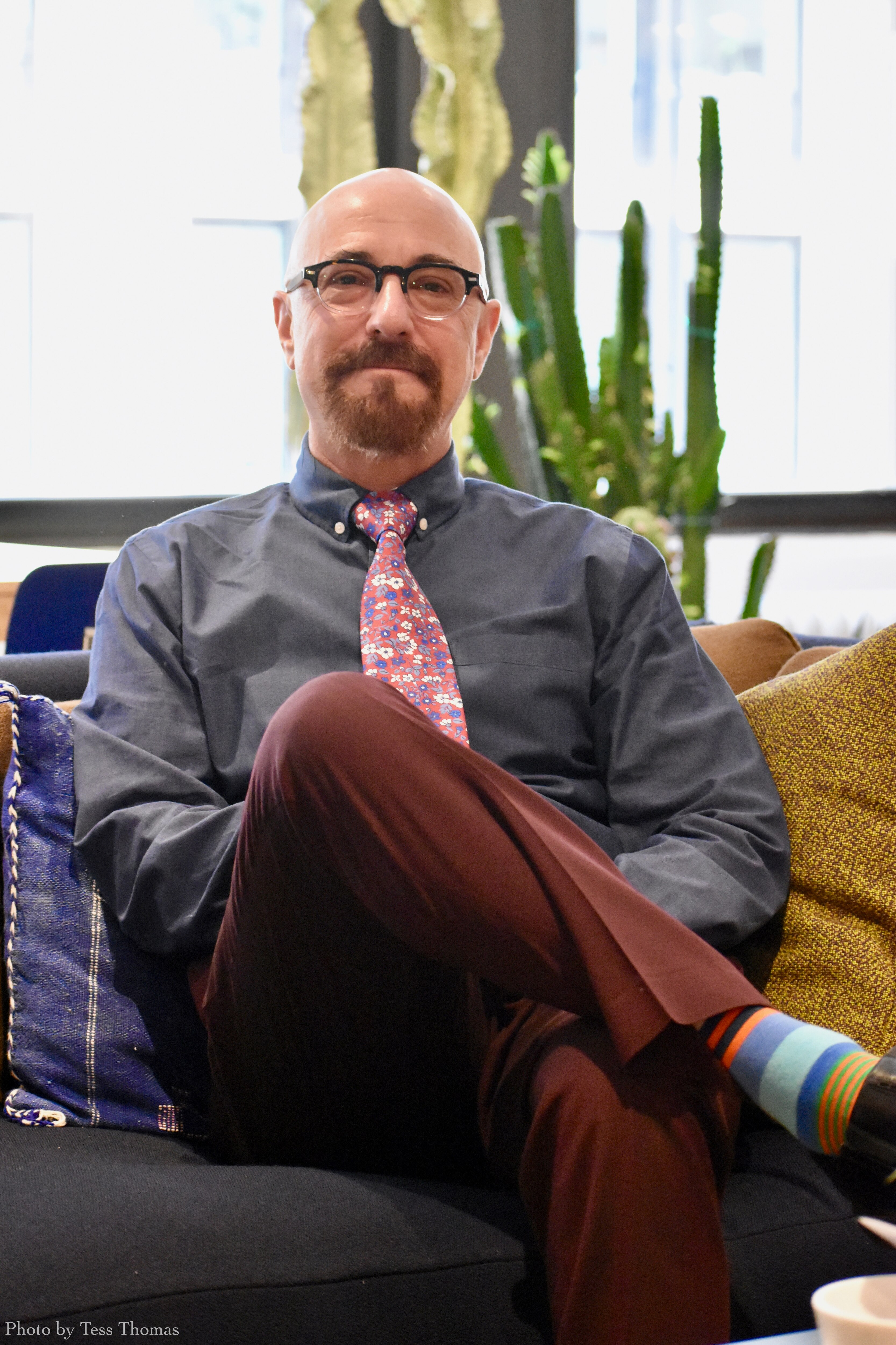 A middle-aged man with a bald head and brown beard sits on a sofa, smiling.