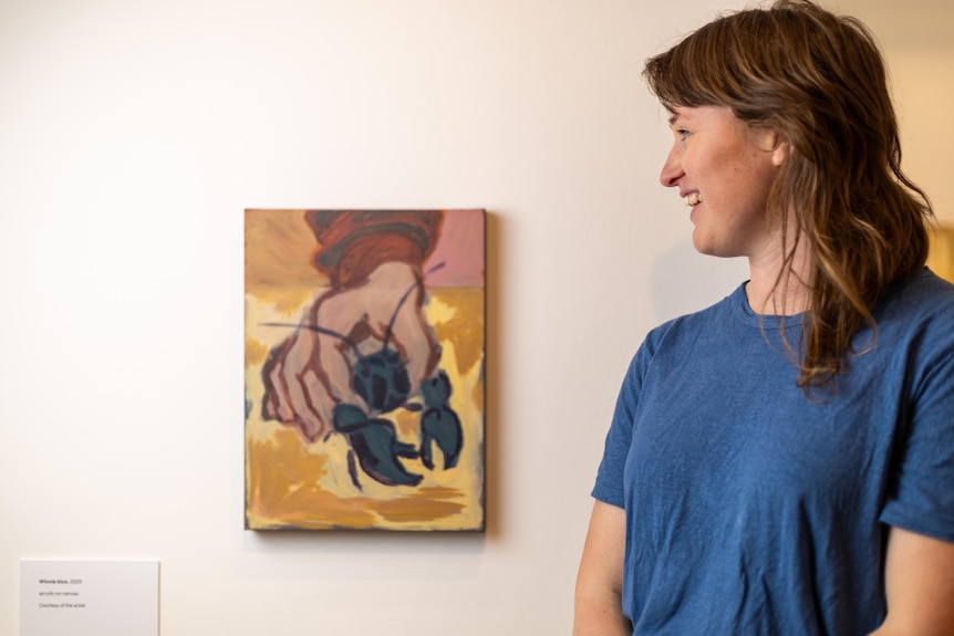 A smiling, dark-haired woman looks at a painting of a yabbie hanging on a wall in an art gallery.