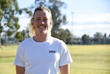 A man with a bleached blonde mullet wearing a white t shirt stands in a green sports oval smiling.