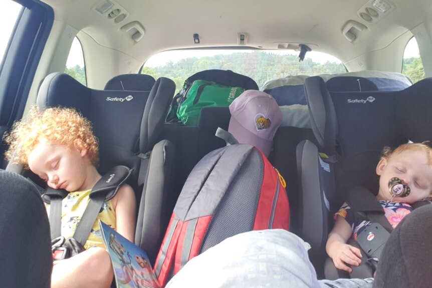 A toddler and baby asleep in child seats in the back of a car packed with stuff