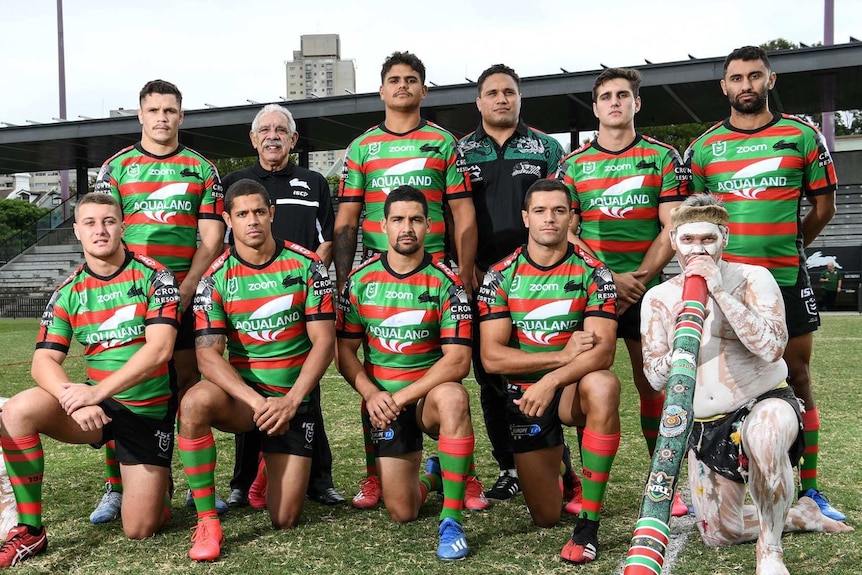 A number of Indigenous players pose for a photo in their jerseys. Two painted Indigenous man with didgeridoos are with them