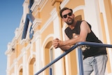 phil jamieson stands on a balcony looking into the distance he wears a tank top and sunglasses.