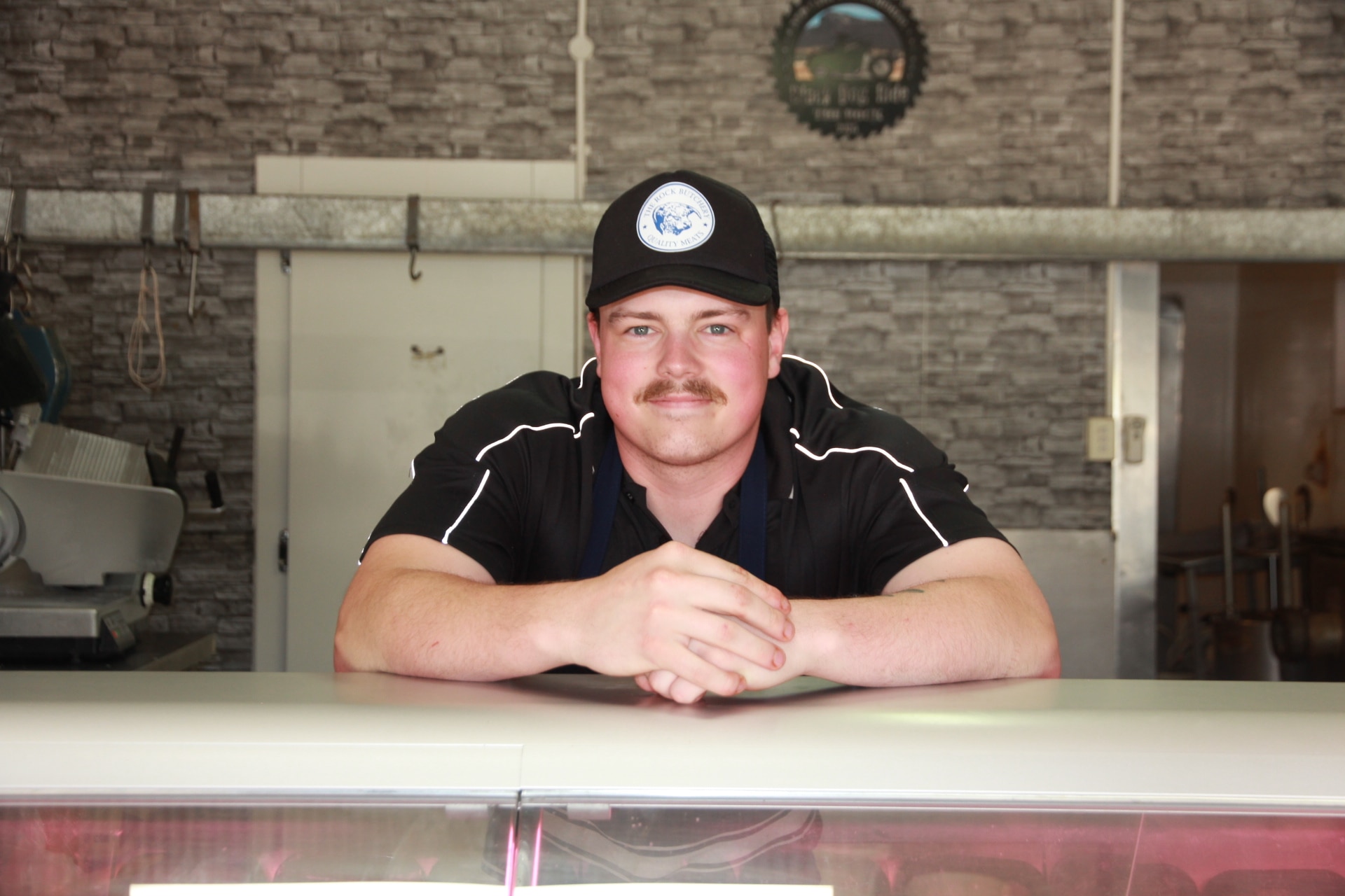 Jovem de camisa preta e chapéu apoiado na vitrine de um açougue