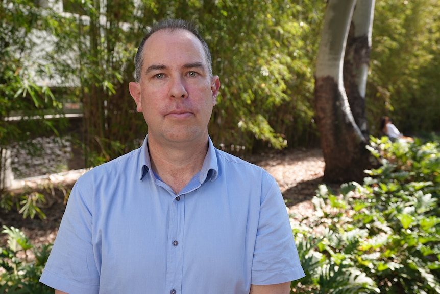 A man looks forwards with forest in the background. 