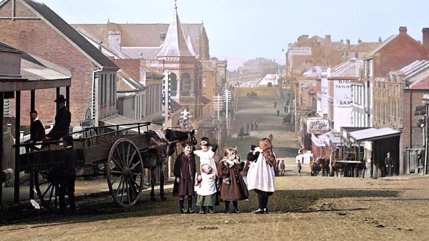 A group of children stand in the road with buildings behind them and a horse and cart beside them.
