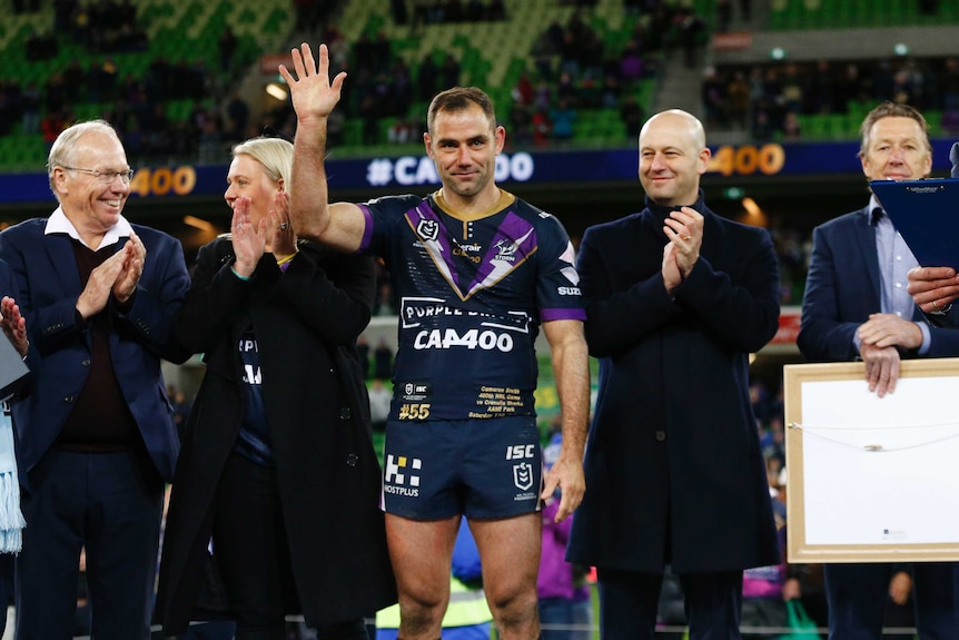 An NRL star stands on a platform next to dignitaries and smiles as he waves to the crowd.