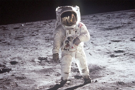 An astronaut in a special suit, walks across the grey surface of the Moon