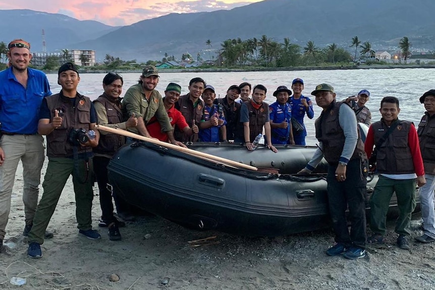 A group of men surround an inflatable boat, water behind them and a mountain in the background.