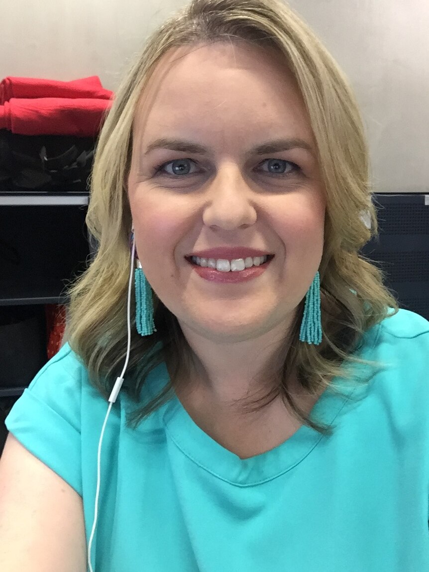 A woman with a turquoise shirt and earrings listens in to a call while working from home. She's smiling.