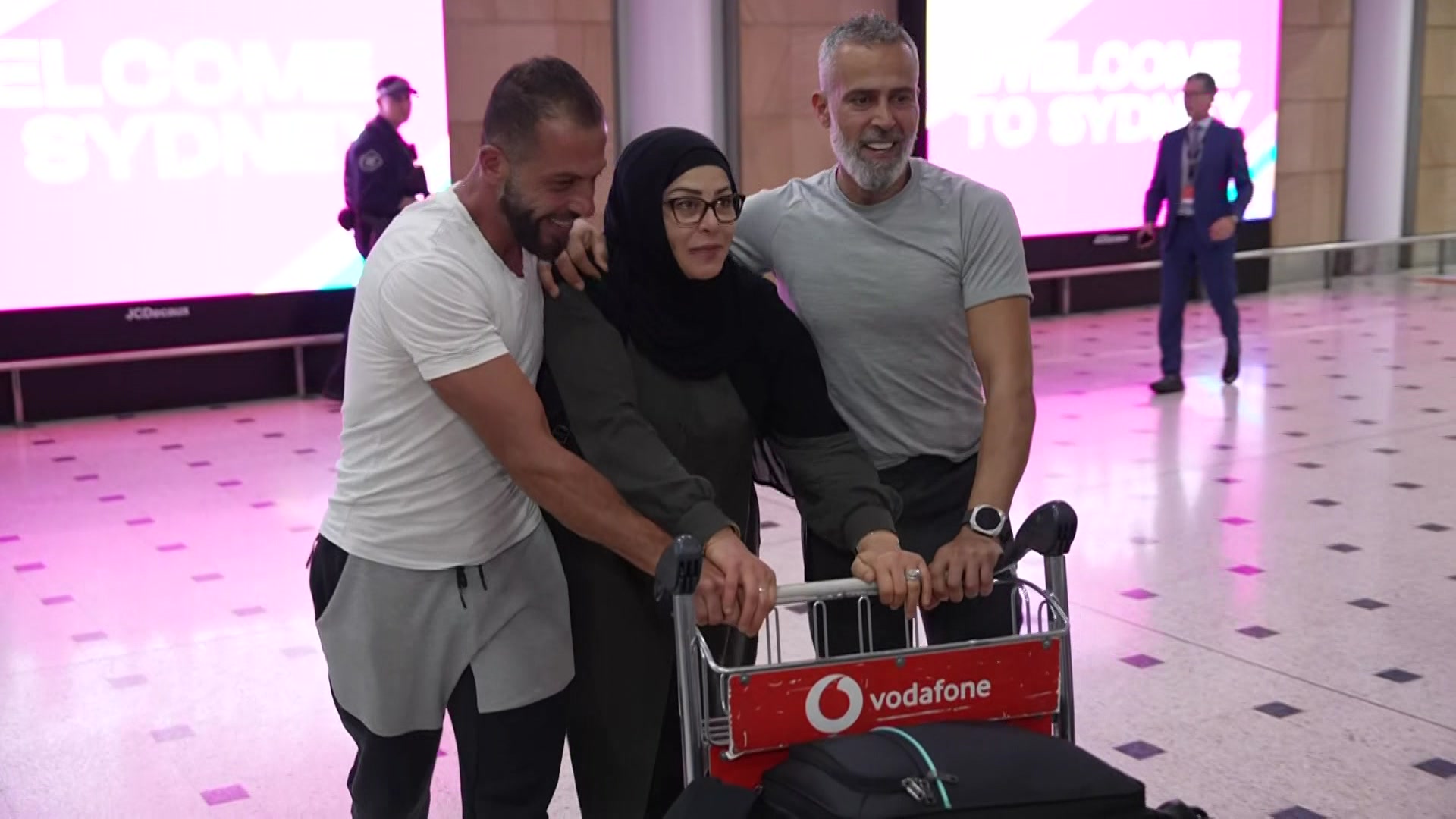 Una mujer que lleva un hijab sonríe mientras dos hombres la ayudan con su carrito de equipaje en el aeropuerto.