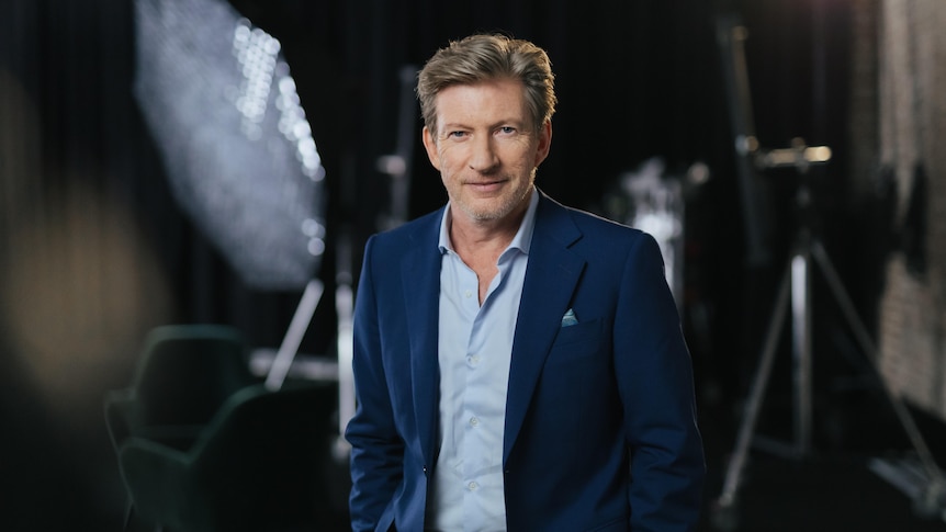 A publicity shot of David who wears a blue suit and light blue shirt, with the background out of focus
