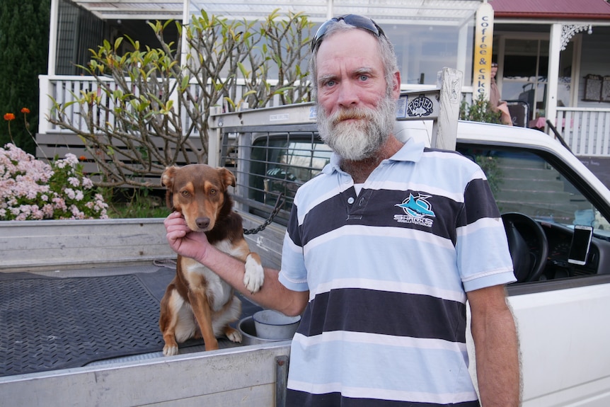 A man in a blue and black striped shirt standing in front of his truck with his dog.