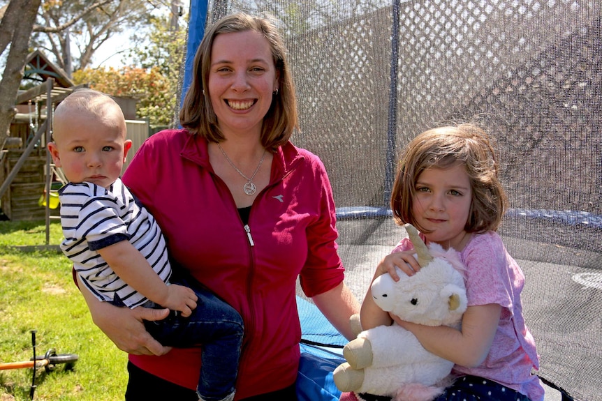 A woman in her backyard, holding her one-year-old son and next to her four-year-old daughter.