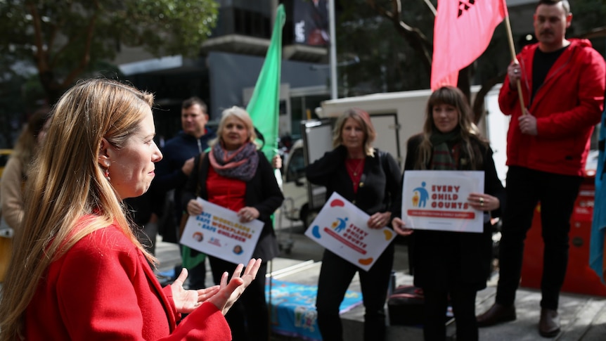 A woman wearing a red jacket addresses a small crows of people holding placards. 