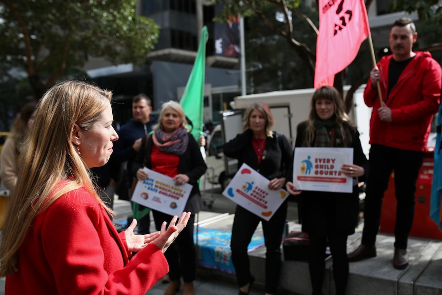 A woman wearing a red jacket addresses a small crows of people holding placards. 