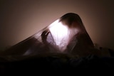 A child's shadow can be seen under a bed sheet, lit by a tablet's backlight.