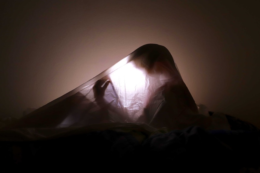 A child's shadow can be seen under a bed sheet, lit by a tablet's backlight.