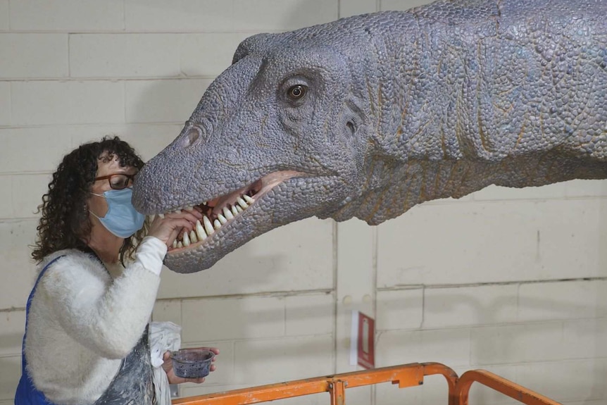 A women is standing and has her hand in the mouth of a giant model dinosaur working on its teeth
