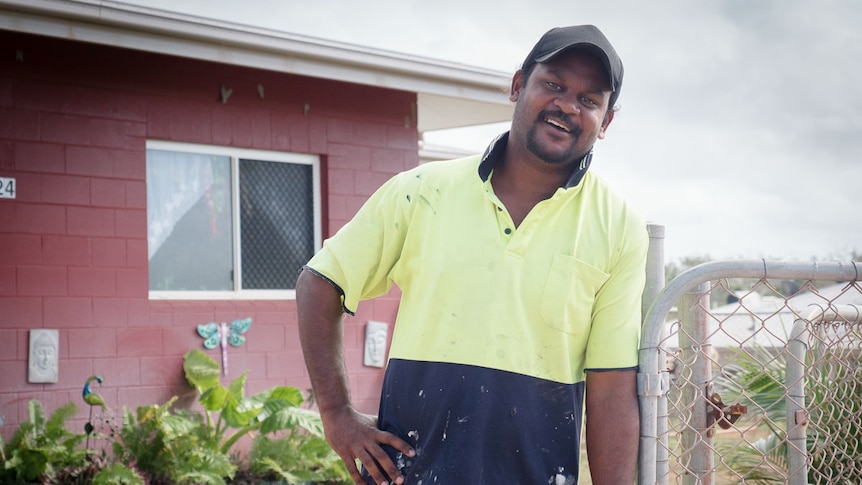 Aboriginal man leans on fence outside of family home.