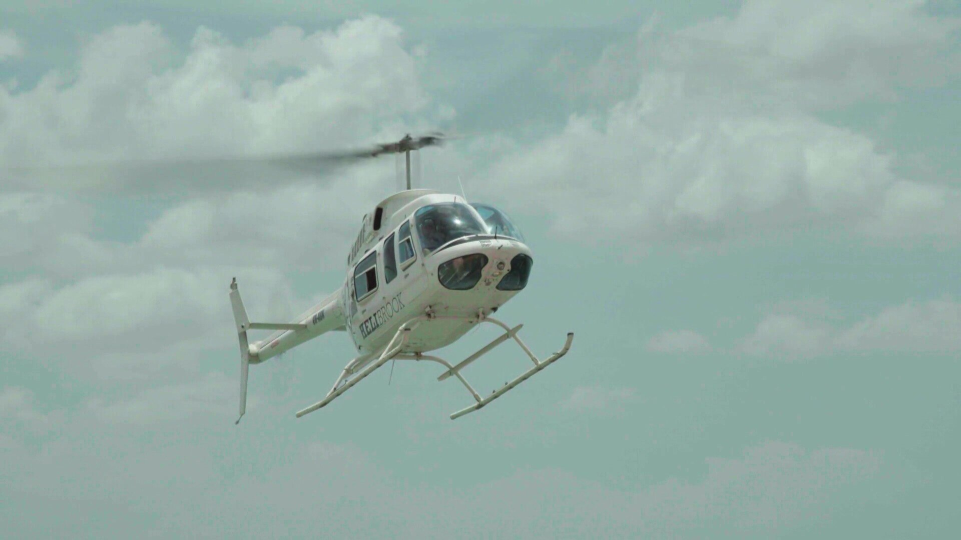 A white helicopter with the word 'Helibrook' on the side, descending to land on the ground.
