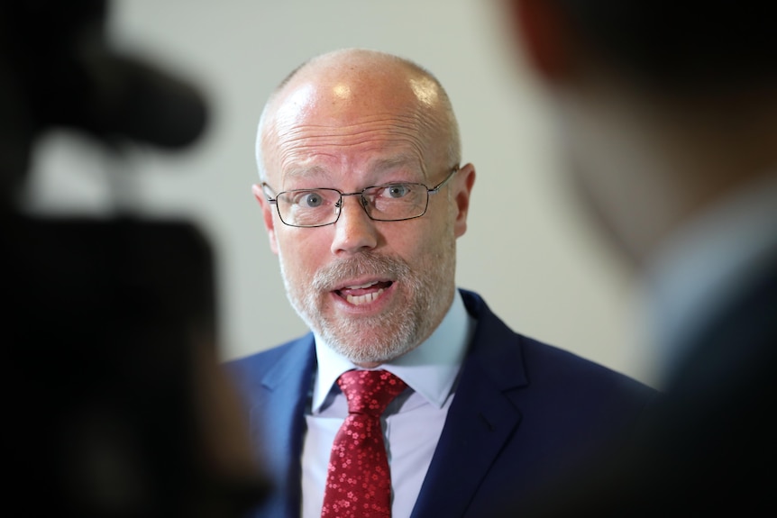 A bald man wearing glasses is speaking to silhouetted figures.