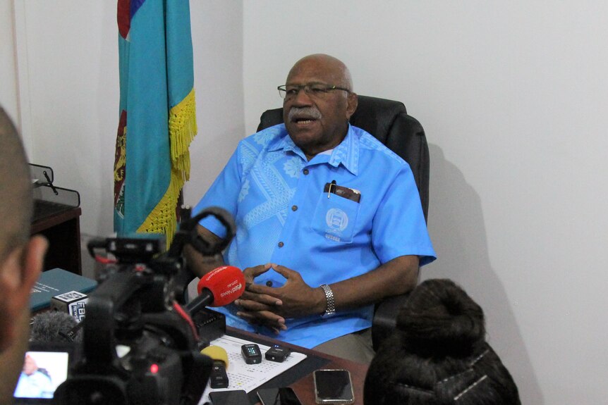 Fijian politician Sitiveni Rabuka sits at his desk surrounded by a media scrum