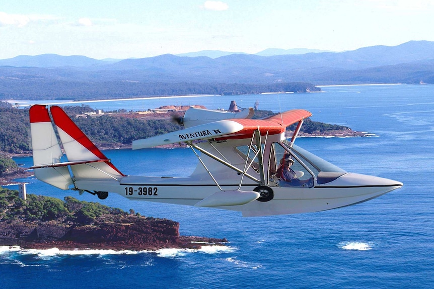 A small white and red plane flies over a rugged coastline on a clear day.