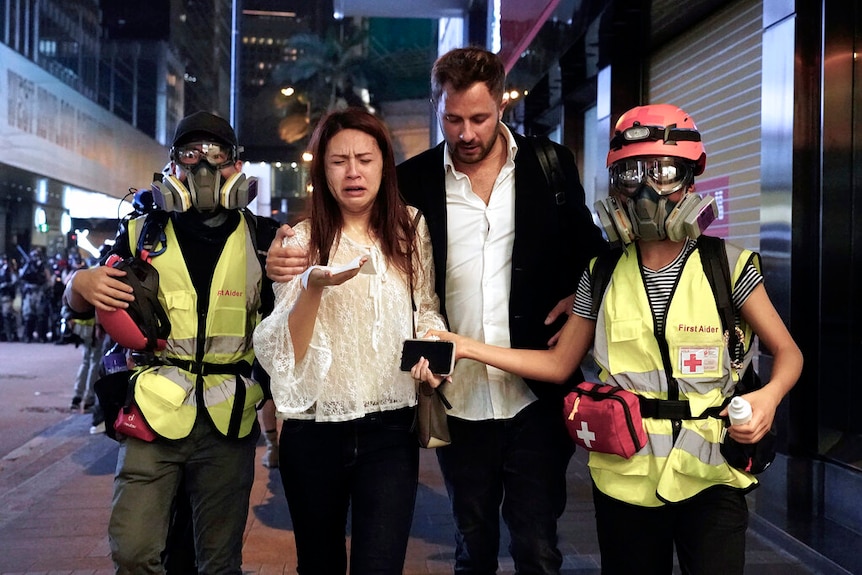You see a couple in smart casual clothes reacting to being struck by tear gas, who are accompanied by two medics.