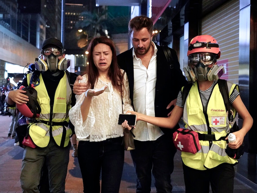 You see a couple in smart casual clothes reacting to being struck by tear gas, who are accompanied by two medics.